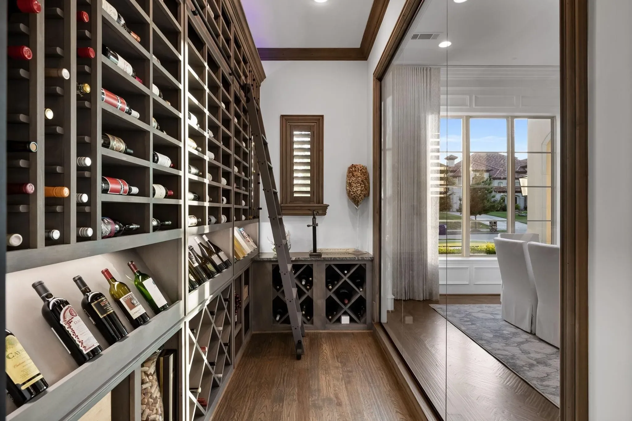Wine room with floor-to-ceiling bottle racks, a rolling library ladder and a glass wall onto the dining room at Craig Ranch, McKinney