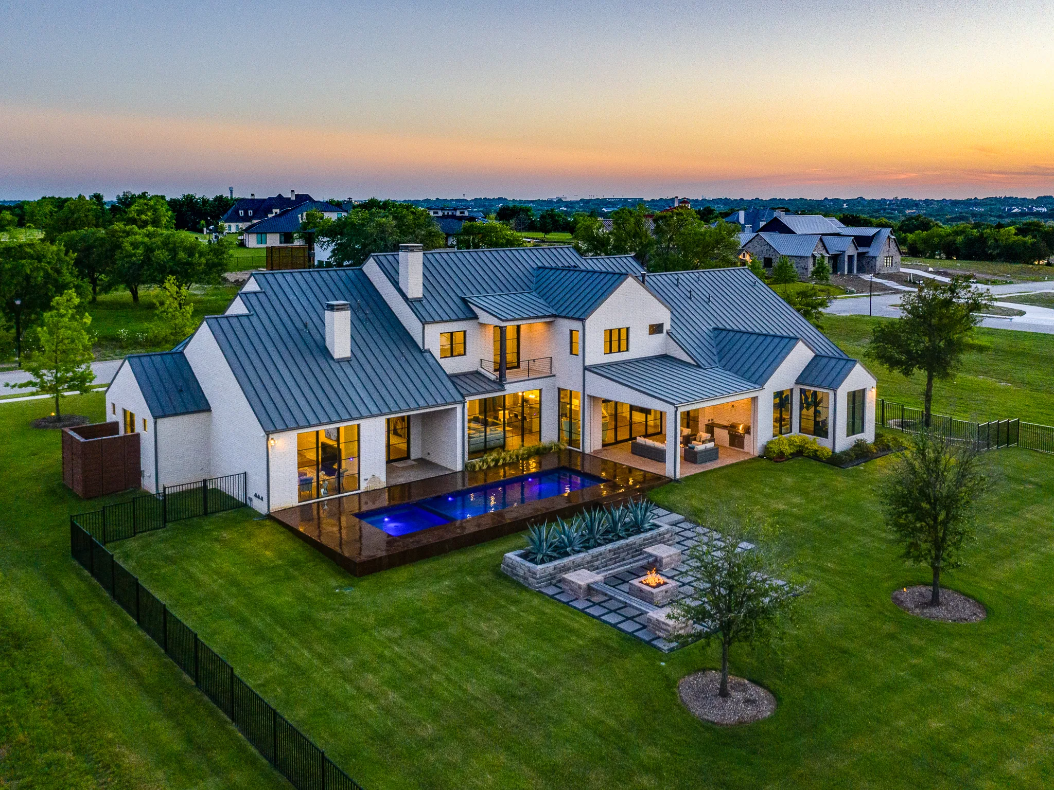 Aerial view at dusk of a white brick home with a metal roof, a lit pool on a wood deck and a fire pit terrace in Ridge Lakes of Heath