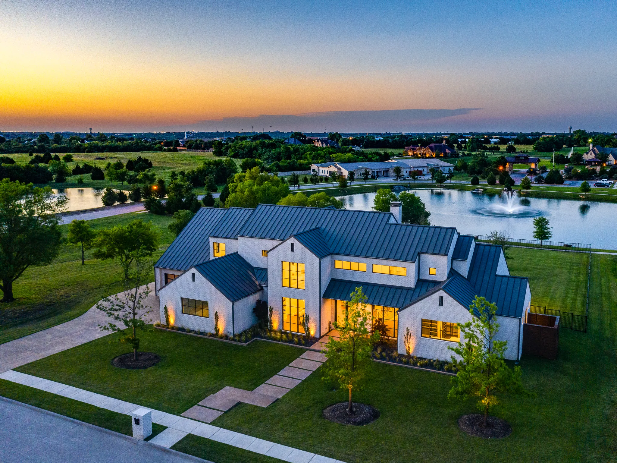 Aerial view at dusk of a finished white brick home with gabled wings and a pond with a fountain behind it in Ridge Lakes of Heath