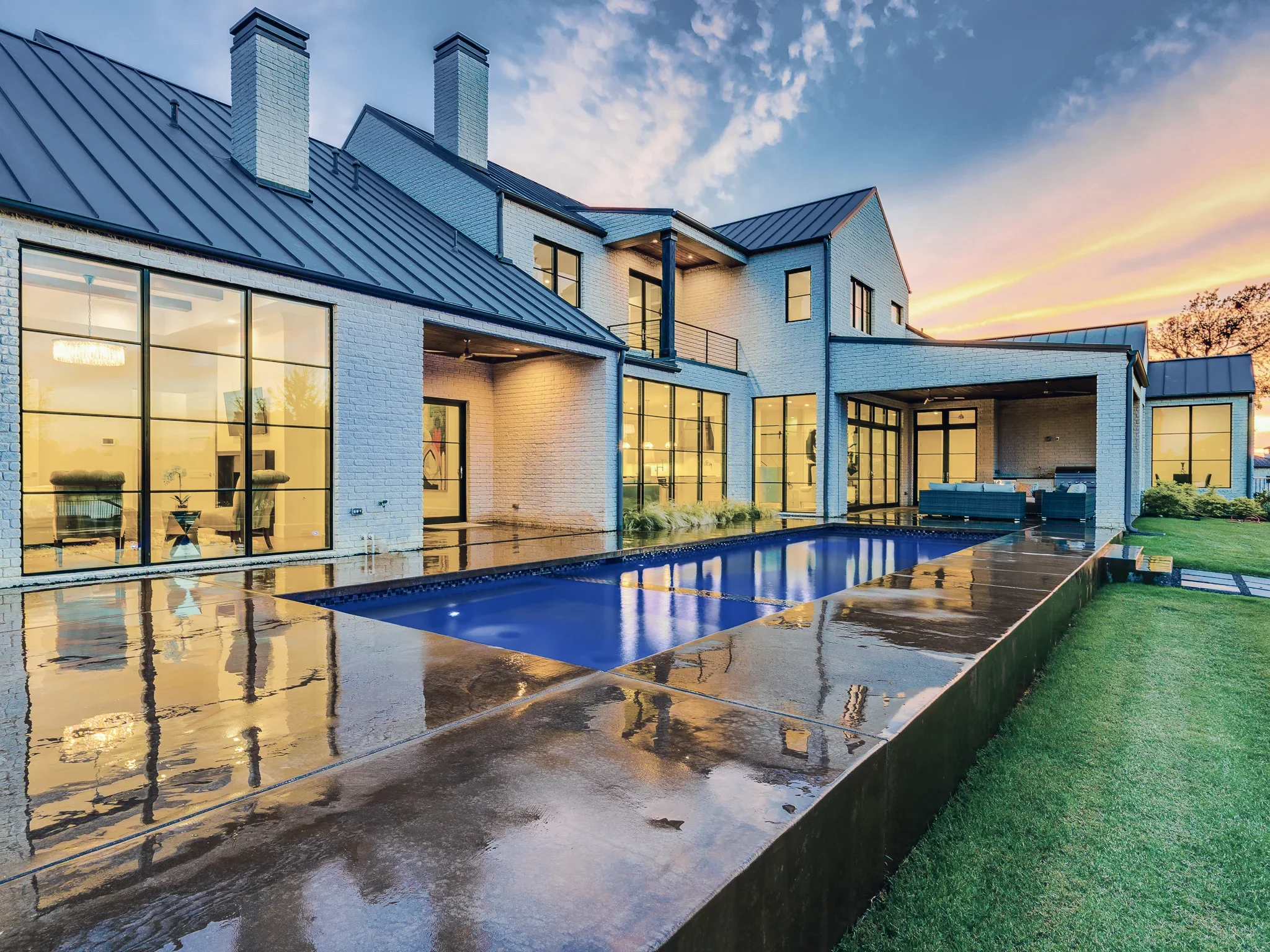 Rear of a white brick home at twilight with a long lit pool, black-framed glass walls and a covered patio reflected in the wet deck in Ridge Lakes of Heath