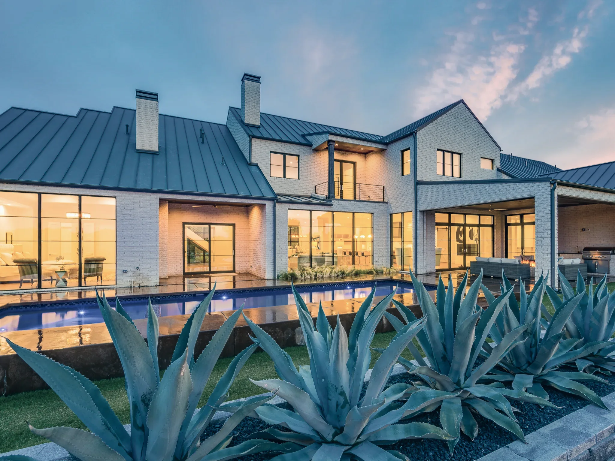 Rear of a white brick modern farmhouse with black-framed glass walls, a standing seam metal roof and agave beside the pool in Ridge Lakes of Heath