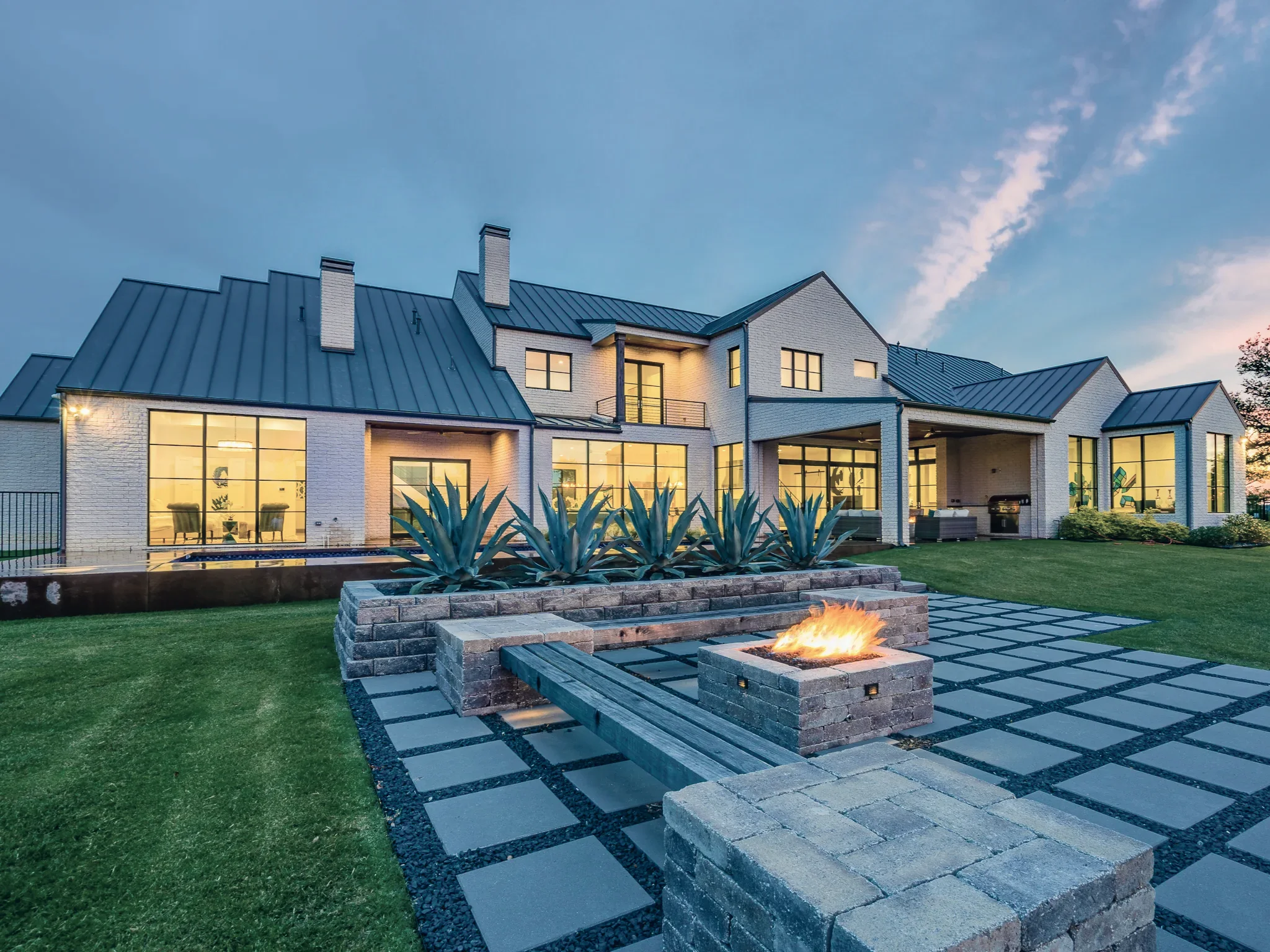 Rear elevation at dusk with a standing seam metal roof, lit windows, agave beds and a stone fire pit burning on the paver terrace in Ridge Lakes of Heath